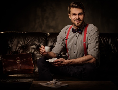 Young handsome old-fashioned bearded man with cup of coffee sitting on comfortable leather sofa on dark background.の写真素材