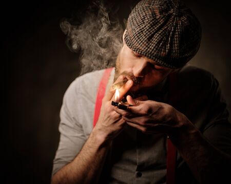 Serious old-fashioned man in tweed hat lighting a cigar on dark background.の写真素材