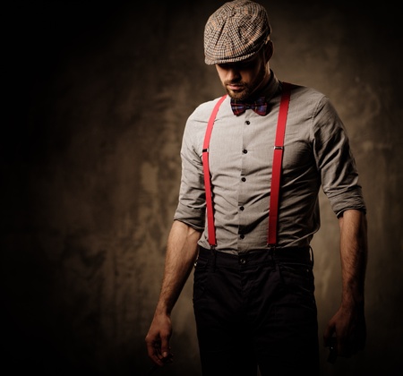 Serious old-fashioned man in tweed hat wearing suspenders and bow tie, posing on dark background.の写真素材
