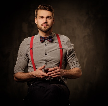 Young handsome old-fashioned bearded man with glass of brandy sitting on dark background.の写真素材