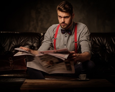 Young handsome old-fashioned bearded man with newspaper sitting on comfortable leather sofa on dark...