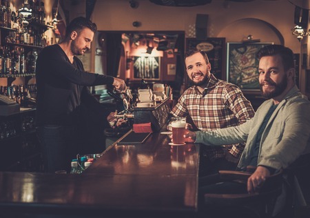 Cheerful old friends drinking draft beer at bar counter in pub.の写真素材