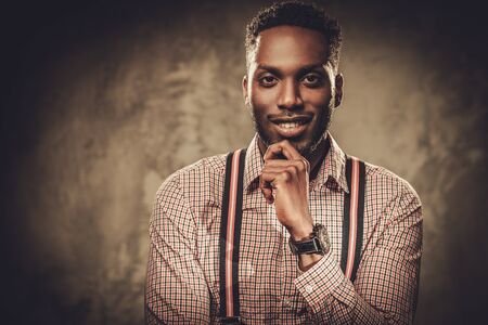 Stylish young black man with suspenders posing on dark background.