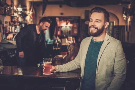 Cheerful stylish man having fun watching a football game on TV and drinking draft beer at bar counter in pub.の写真素材