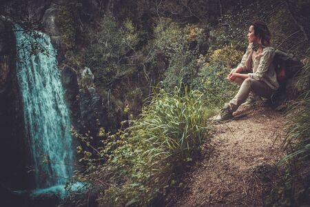 Beautiful woman hiker sitting near waterfall in deep forest.の写真素材