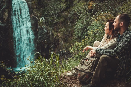 Cheerful couple hikers sitting near waterfall in deep forest.の写真素材