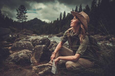 Smiling woman hiker, sitting with map in her hands, near wild mountain river.の写真素材