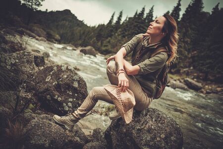 Beautiful woman hiker sitting on the stone near wild mountain river.