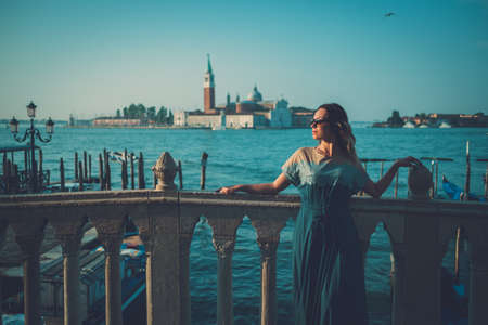 Beautiful well-dressed woman standing near San Marco square with gondolas and Saint George island on the background.の写真素材
