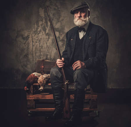 Senior hunter with a shotgun in a traditional shooting clothing, sitting on a dark background.の写真素材