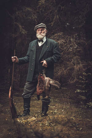 Senior hunter with a shotgun and pheasants in a traditional shooting clothing on a dark forest background.の写真素材