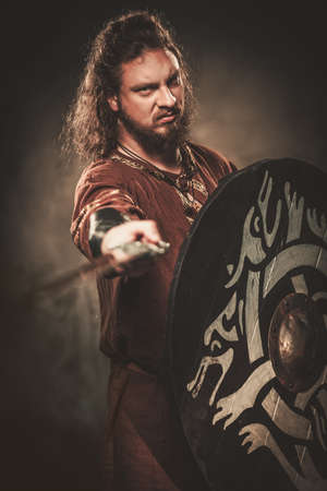 Serious viking with sword in a traditional warrior clothes, posing on a dark background.