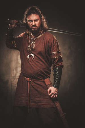 Serious viking with sword in a traditional warrior clothes, posing on a dark background.の写真素材