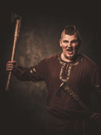 Angry viking with ax in a traditional warrior clothes, posing on a dark background.の写真素材