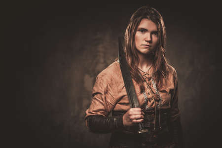 Serious viking with sword in a traditional warrior clothes, posing on a dark background.の写真素材
