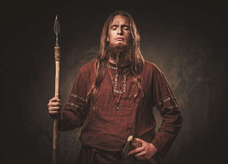 Serious viking with a spear in a traditional warrior clothes on a dark background.