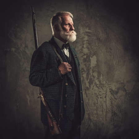 Senior hunter with a shotgun in a traditional shooting clothing on a dark background.の写真素材