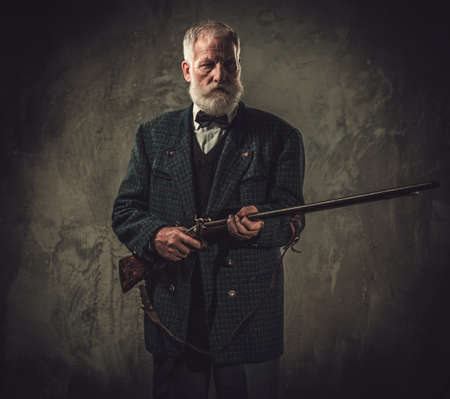 Senior hunter with a shotgun in a traditional shooting clothing on a dark background.の写真素材