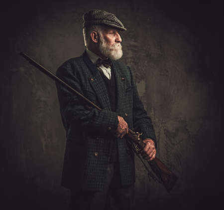 Senior hunter with a shotgun in a traditional shooting clothing on a dark background.の写真素材