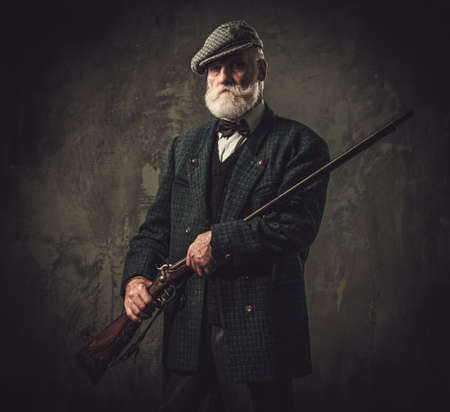 Senior hunter with a shotgun in a traditional shooting clothing on a dark background.の写真素材