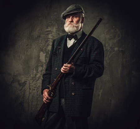 Senior hunter with a shotgun in a traditional shooting clothing on a dark background.の写真素材