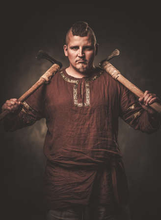Angry viking with axes in a traditional warrior clothes, posing on a dark background.の写真素材