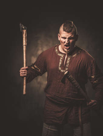 Angry viking with axes in a traditional warrior clothes, posing on a dark background.の写真素材