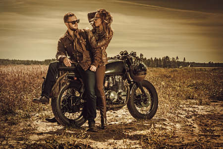 Young, stylish cafe racer couple on the vintage custom motorcycles in a field.の写真素材