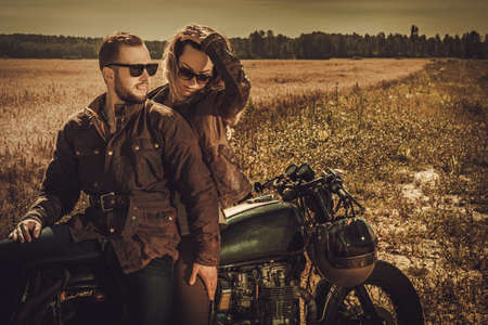 Young, stylish cafe racer couple on the vintage custom motorcycles in a field.