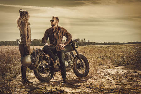 Young, stylish cafe racer couple on the vintage custom motorcycles in a field.