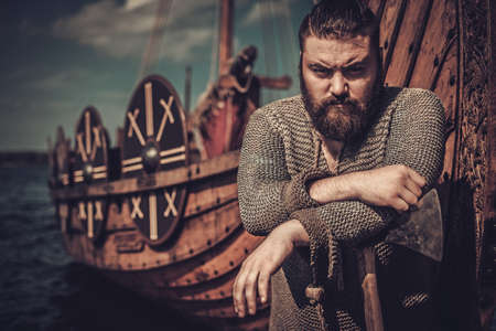 Serious viking warrior with axe standing near Drakkar on the seashore.