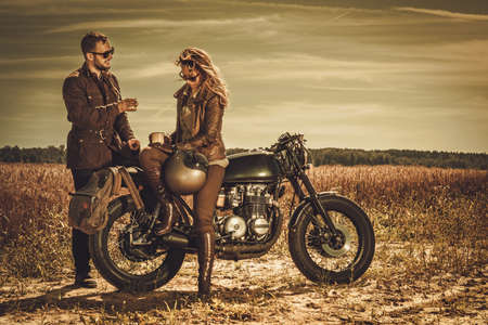 Young, stylish cafe racer couple on the vintage custom motorcycles in a field.の写真素材