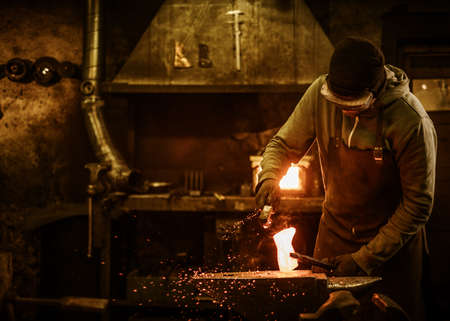 The blacksmith with brush handles the molten metal on the anvil in smithy.