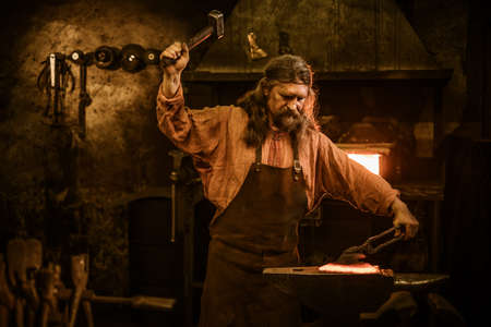 Senior blacksmith forging the molten metal on the anvil in smithy.