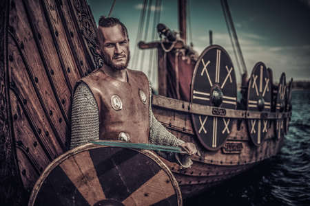 Viking warrior with sword and shield standing near Drakkar on the seashore.