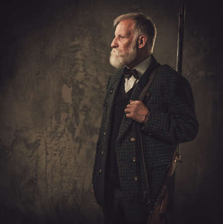 Senior hunter with a shotgun in a traditional shooting clothing, posing on a dark background.