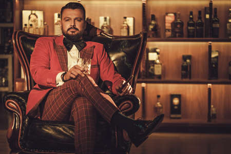 Extravagant stylish man with whisky glass sitting on armchair in gentleman club.の写真素材