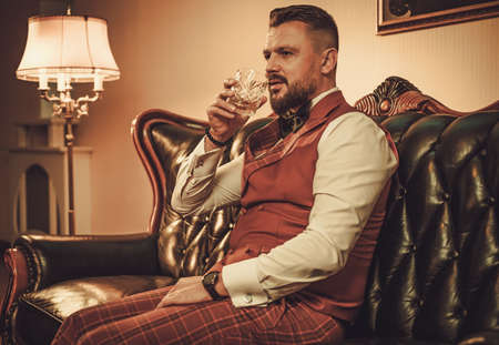 Extravagant stylish man with glass of whisky sitting on classic leather sofa in gentleman club.の写真素材