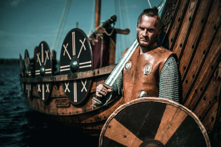 Serious viking warrior with sword and shield standing near Drakkar on the seashore.