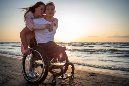Handicapped man in wheelchair and his girlfriend on a beach at sunsetの写真素材