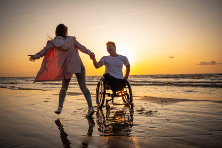 Handicapped man in wheelchair and his girlfriend on a beach at sunsetの写真素材