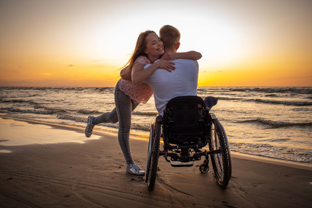 Handicapped man in wheelchair and his girlfriend on a beach at sunsetの写真素材