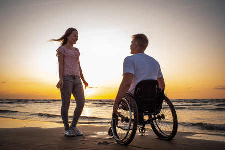 Handicapped man in wheelchair and his girlfriend on a beach at sunsetの写真素材