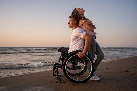 Handicapped man in wheelchair and his girlfriend on a beach at sunsetの写真素材