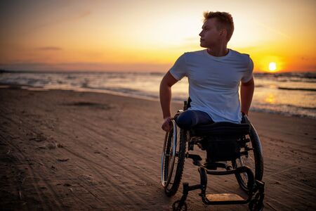 Handicapped man in wheelchair and his girlfriend alone on a beach at sunsetの写真素材