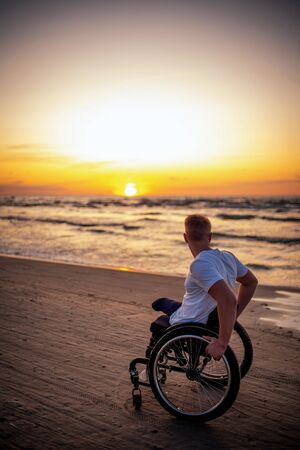 Handicapped man in wheelchair and his girlfriend alone on a beach at sunset.の写真素材