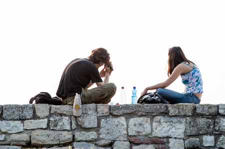 Young Charming couple sitting Side By Side on stonewall, relaxing and drinking, isolated on white (from the rear).の写真素材
