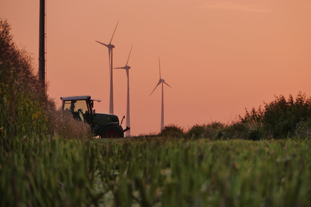 A Tractor crossing the scenery of a field of mowed down straws of wheat and some wind tubines in the background under a red skyの写真素材