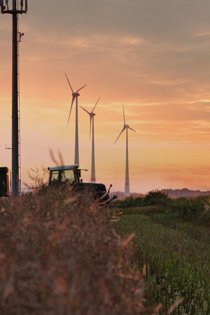 A Tractor crossing the idyllic scenery of a mowed down field and some wind tubines in the background while the sun sets in the far distanceの写真素材