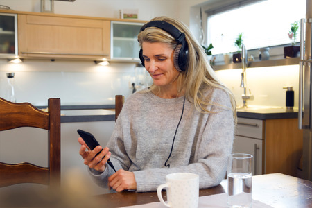 Portrait of relaxed middle-aged woman with headphones sitting at kitchen table and listening music on smartphone. Cup of coffee and glass of water in the foregroundの写真素材